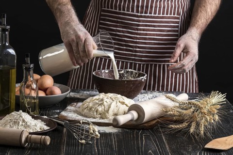 Baker in uniform pouring milk from plastic bottle into bowl. A handful of flour with egg on a