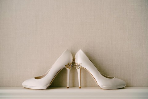 White women's shoes with high pointed heels, with a pattern of flowers, on a gray background.