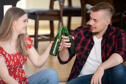 Smiling young people drink beer at a house party. They tap green bottles and look at each other. A