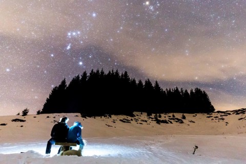 The Milky Way over the winter mountain landscape with pine trees. Two people sitting in the