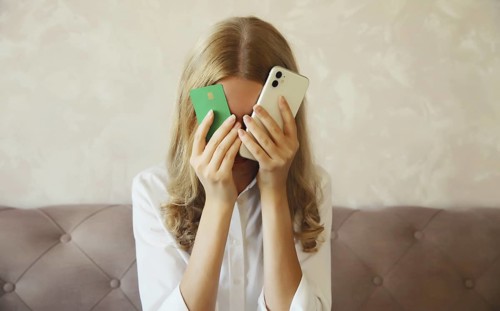 Desperate upset young woman holding mobile phone and bank card touching her head and covering face,