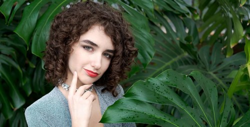 Woman with curly hair is standing in front of a green plant. She is wearing a blue dress and a