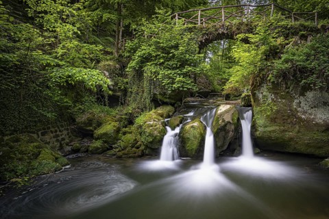 Three small waterfalls under a wooden bridge, surrounded by green vegetation, Mullerthal Region,