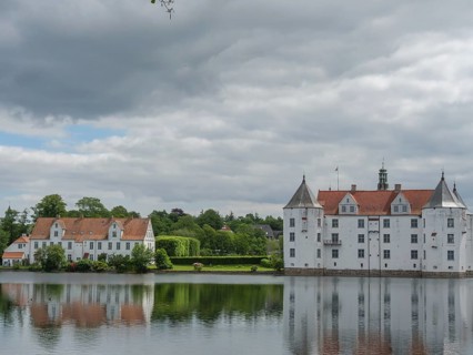 A historic castle with white facades and red roofs is reflected in a calm body of water under a