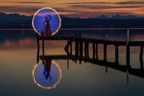 Woman in a red dress on a jetty at sunset on a lake, standing in front of a circle of light created