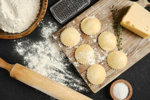 Fresh pasta dough is rolled out on a wooden board surrounded by flour, cheese, and herbs. Round