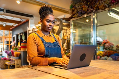Young florist managing online orders using laptop in her flower shop, surrounded by colorful