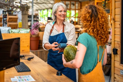 Smiling greengrocer holding pos terminal while customer paying with contactless payment for