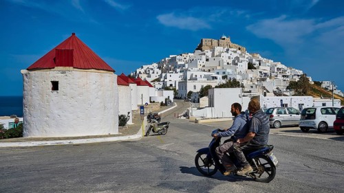Two people on motorbike in front of white village and windmills on hill, Querini Castle, Chora,