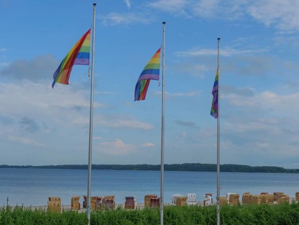 Three rainbow flags waving on the beach above a row of beach chairs, calm water in the background,