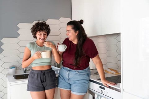 A lesbian couple stands in a cozy kitchen, sipping morning coffee. The modern decor and soft