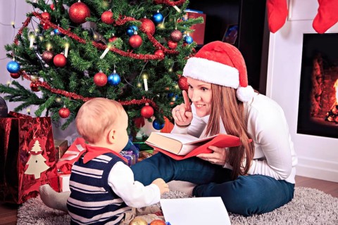 Toddler boy and his mother reading a book in front of christmas tree