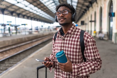 Traveler with headphones and backpack holding smartphone and reusable coffee cup is waiting for