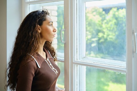 A young woman with long, curly hair is looking out a large window. Bright sunlight fills the room,