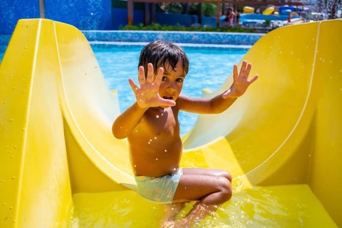 Happy child sliding down yellow water slide in amusement park, having fun during summer vacation