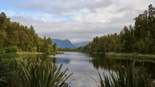 A tranquil lake surrounded by lush vegetation and mountains, with a slightly cloudy sky, summer,