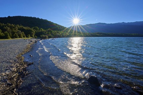 A tranquil lake with sunbeams hitting the water and shore, surrounded by forested mountains under
