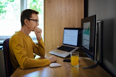 A middle-aged man sits at a wooden desk in a home office, focused on a computer screen displaying