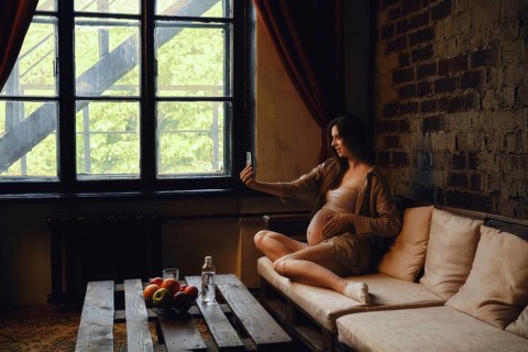 A pregnant woman sits on a comfortable couch at home, casually taking a selfie with her phone.