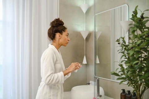 In a contemporary bathroom, a woman dressed in a white robe stands before a mirror, carefully
