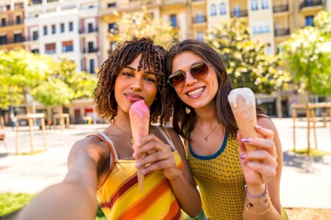 Two happy female friends enjoying ice cream cones on a sunny day in the city, taking a selfie to