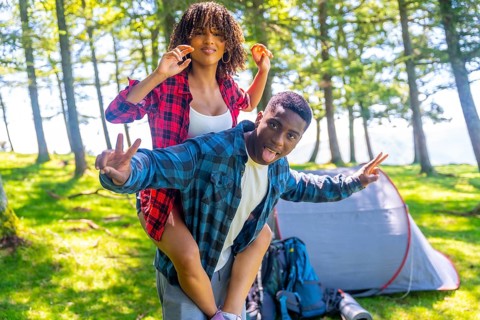 Young couple having fun piggybacking and making funny faces near their tent in a forest campground