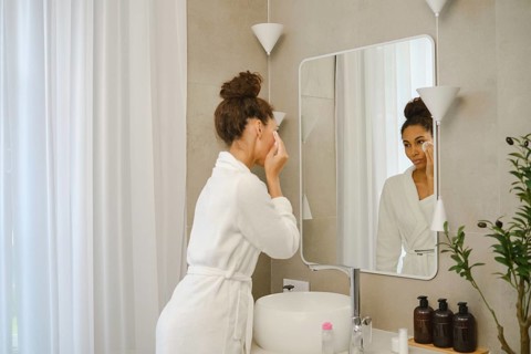 A woman in a white bathrobe is applying skincare products in a contemporary bathroom. She stands