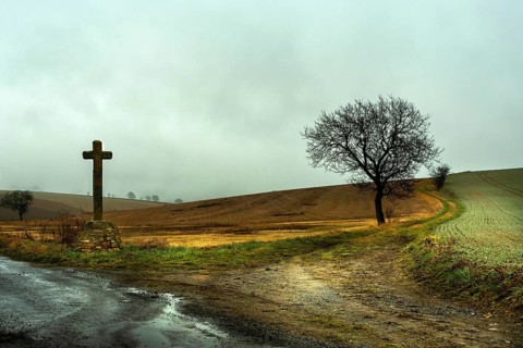 A solitary wooden cross stands beside a muddy dirt road, surrounded by rolling hills under a gloomy