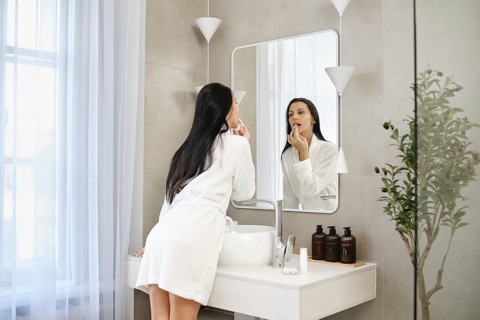 A woman in cotton robe is washing off lipstick in a bathroom, standing in front of a large mirror