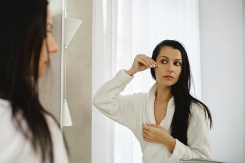 A woman in a white robe looks into a mirror while applying a beauty treatment to her under eye skin