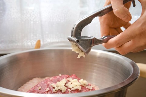 A hand is using a garlic press to add freshly minced garlic to a bowl with ground meat. This