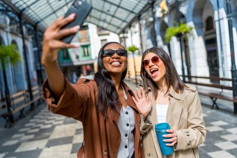 Two cheerful businesswomen taking a selfie with a smartphone and waving at the camera while walking