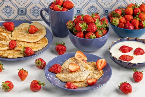 Pancakes sprinkled with icing sugar and fresh strawberries on plates next to yoghurt, blue crockery
