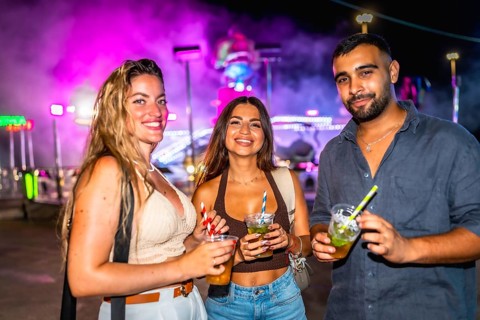 Three friends are holding cocktails while enjoying the vibrant nightlife at an amusement park,