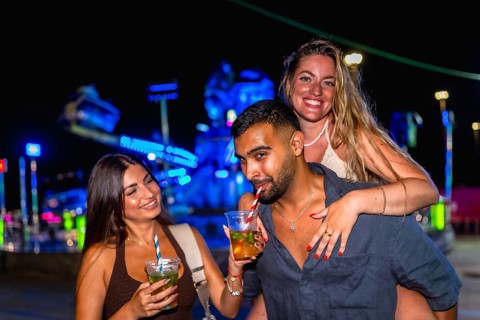 Three cheerful friends enjoying refreshing cocktails at an amusement park, illuminated by vibrant
