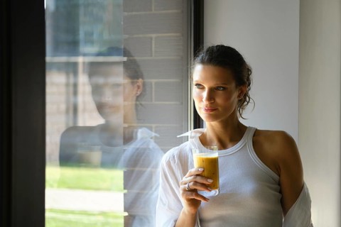 In a stylish, well-lit room, a woman holds a glass of fresh juice. She gazes thoughtfully out the