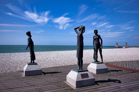 Bronze sculpture La Heure du Bain by Dominique Denry on the beach, sky, blue, behind it beacon Feu
