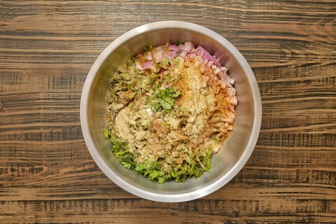 Overhead view of a bowl with chopped herbs, diced onions, and various spices added to minced meat