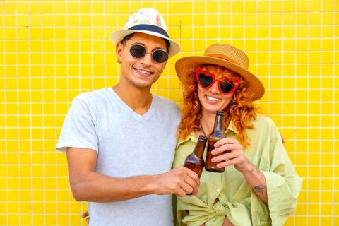 Happy couple toasting with beer bottles while enjoying a summer day against a vibrant yellow tiled