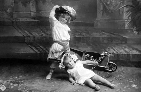 Child falls out of wheelbarrow, 1910, Two children playing in the garden, one with a wreath of