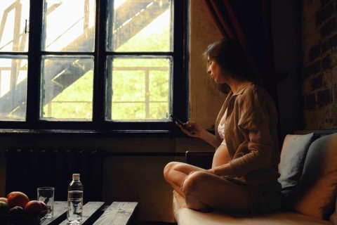 A pregnant woman sits cross-legged on a couch in a cozy room, looking out of the window. She is