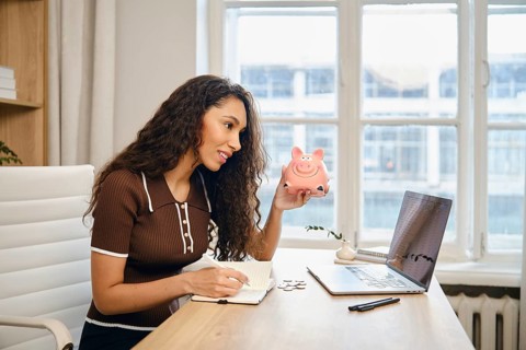 A young woman is actively budgeting at her desk, holding a pink piggy bank in one hand while