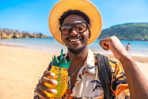 Young man smiling and raising fist while holding a pineapple shaped cocktail on a sunny beach,