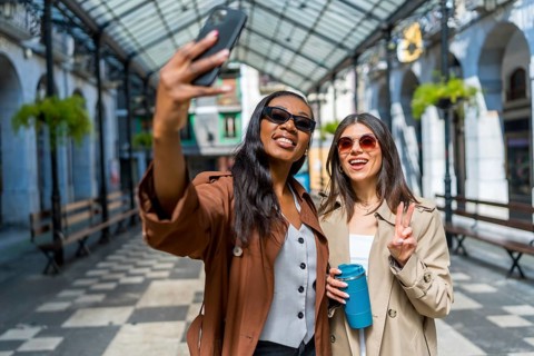 Two female friends taking a selfie in a covered walkway in a city center, one is holding a reusable