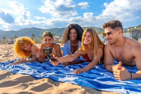 Five young friends lying on a striped towel on a sandy beach, taking a selfie with an action camera
