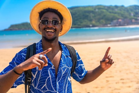 Happy black tourist smiling widely while pointing both index fingers up at a beautiful beach,