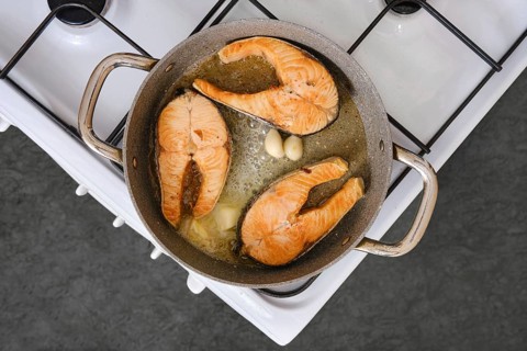 Salmon fillets are being cooked in a pan with garlic on a stovetop. The kitchen is well-lit and the