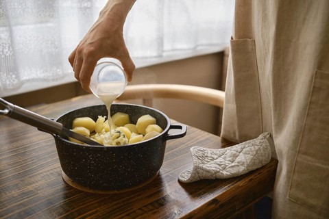A hand pours warm milk onto a pot filled with boiled potatoes on a wooden table. The process of