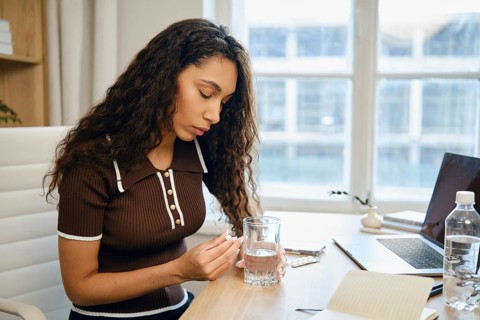 A woman with curly hair sits at a desk in a contemporary office, holding a pill and a glass of