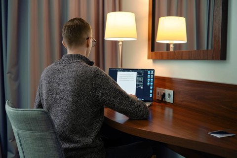 A person sits at a wooden desk in a hotel room, focused on his laptop screen. Soft lamp light